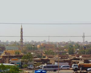 The image displays a busy roadway with numerous cars and vans colored white. Beyond the roadway, lines of dilapidated homes and buildings can be see and the calltower of a mosque can be seen on the lefthand side of the image. The image overall has a gold saturation. It is supposed to depict a city in Sudan. The sky is cloudless and some trees can be seen in the distance.