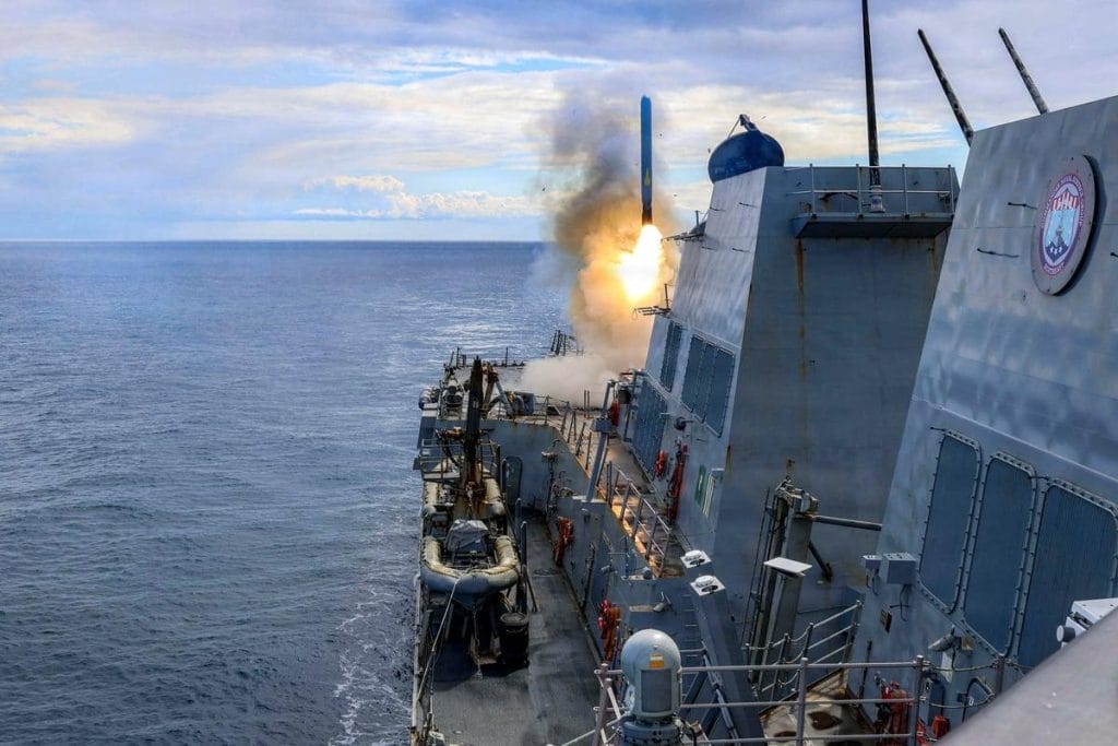 The picture depicts a corner of a large ship sailing on the ocean toward an empty horizon. A missile is being launched from the front of the ship.