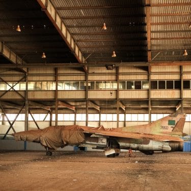 The image depicts a yellow-shaded airplane hanger with a small jet in the middle of the screen. The front third of the jet appears to have fabric placed on top of it.